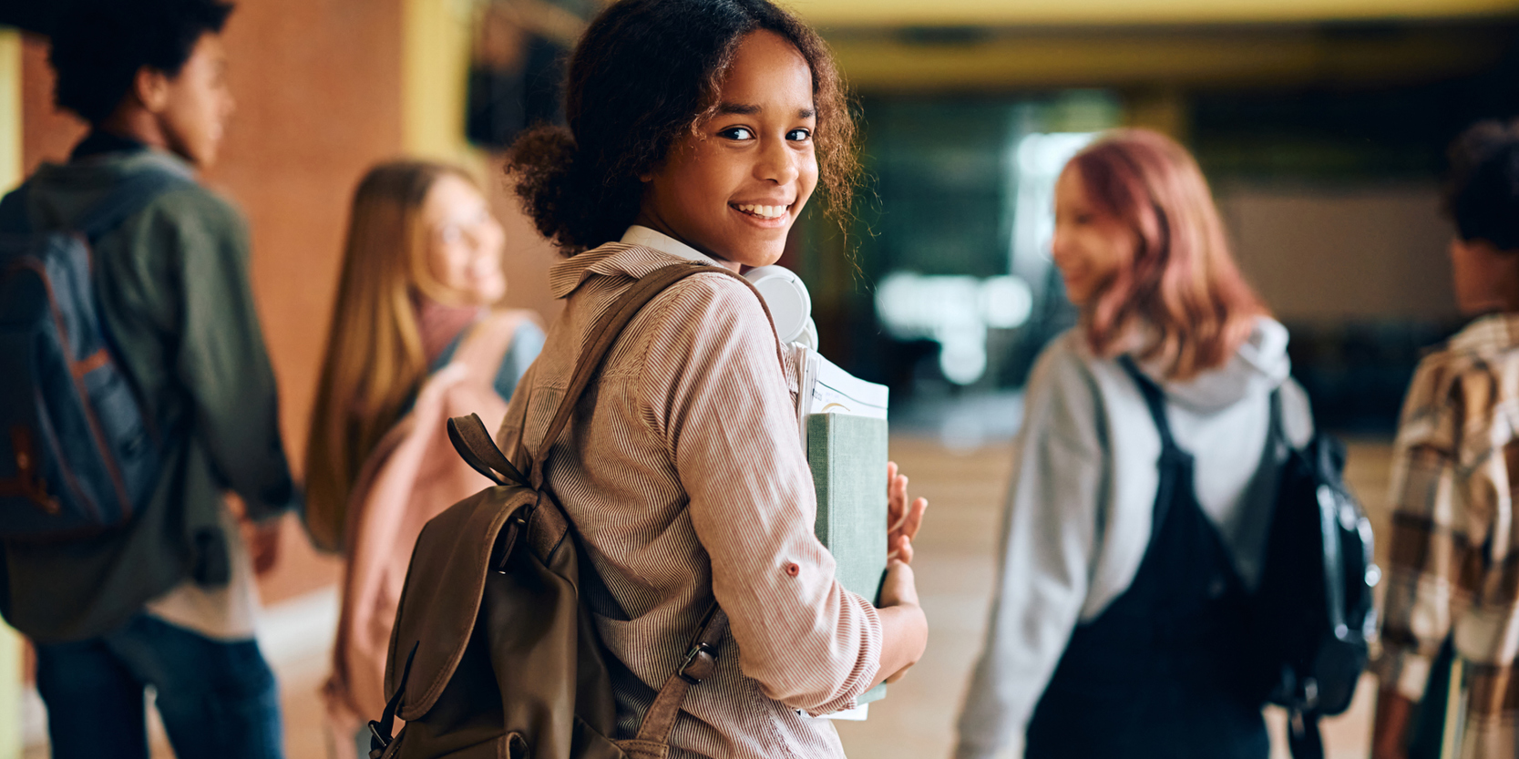 Girl in a school hallway, wearing a backpack and holding textbooks, smiling back at camera